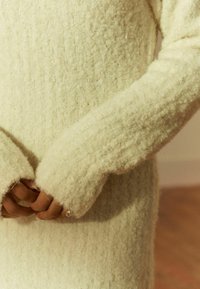 Texturierter, cremefarbener Pullover mit langen, weiten Ärmeln. Der Stoff ist weich und flauschig und weist ein Rippenmuster auf.
