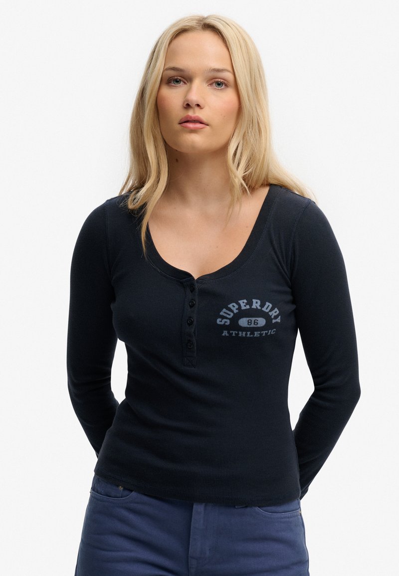 Blonde vrouw met los haar, gekleed in een aansluitend zwart henleyshirt met lange mouwen en de tekst "Superdry Athletic", staand met de handen achter de rug.