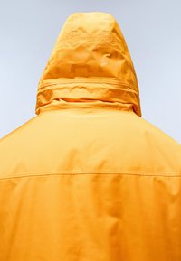 Žlutá nepromokavá bunda s texturovanou kapucí. Hladký materiál, jednoduchý design a švy na ramenou a zádech.