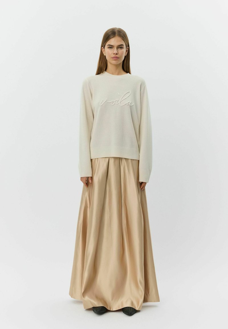 Femme portant un pull crème avec le texte « voilà » et une longue jupe beige brillante, debout devant un fond blanc uni.