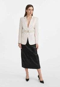 Biała marynarka z paskiem w talii, z klapami, noszona na czarnej skórzanej spódnicy midi. Modelka stoi w czarnych szpiczastych szpilkach.