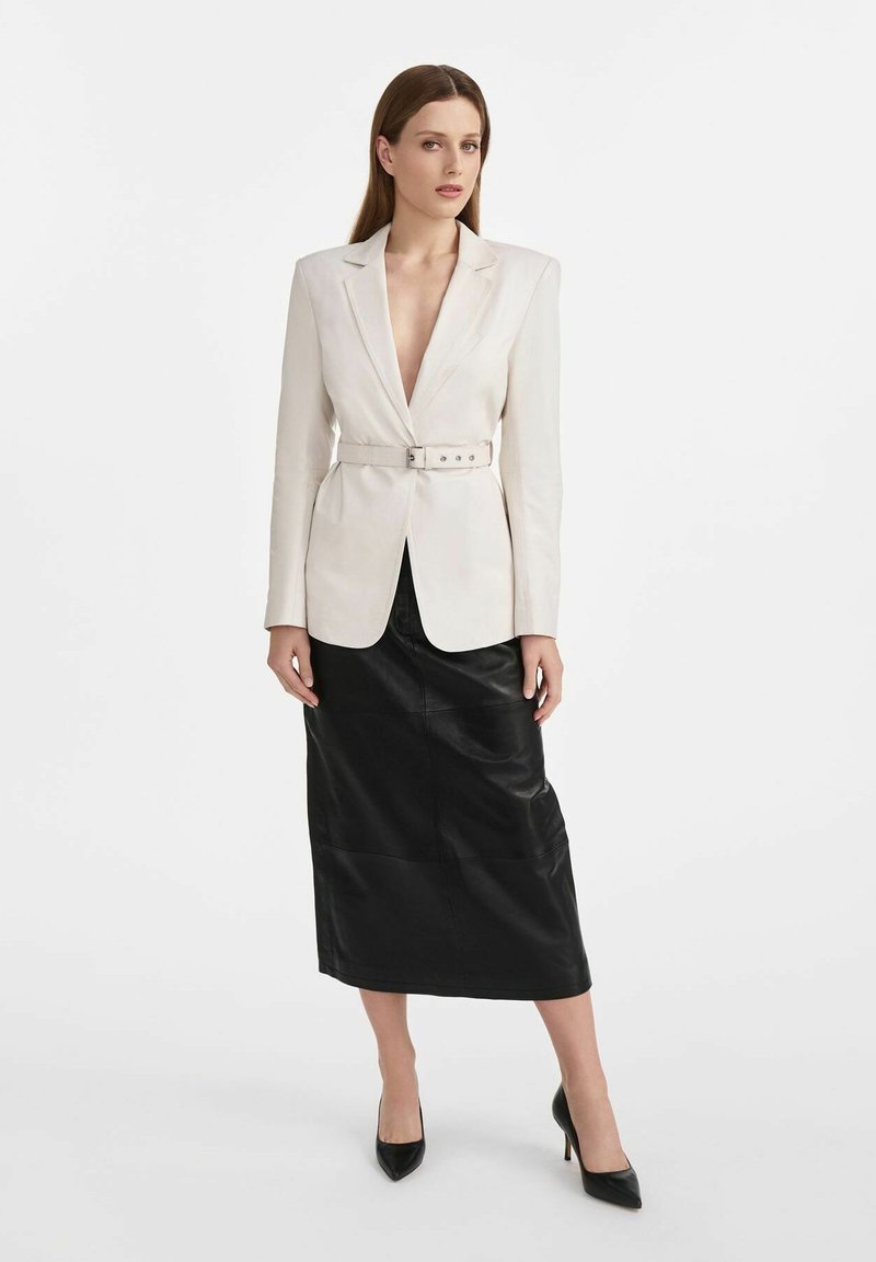 Biała marynarka z paskiem w talii, z klapami, noszona na czarnej skórzanej spódnicy midi. Modelka stoi w czarnych szpiczastych szpilkach.