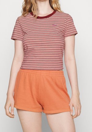 Frau trägt ein rot-weiß gestreiftes Levi’s T-Shirt und orangefarbene Shorts mit elastischem Bund, steht mit entspannt an den Seiten hängenden Armen.