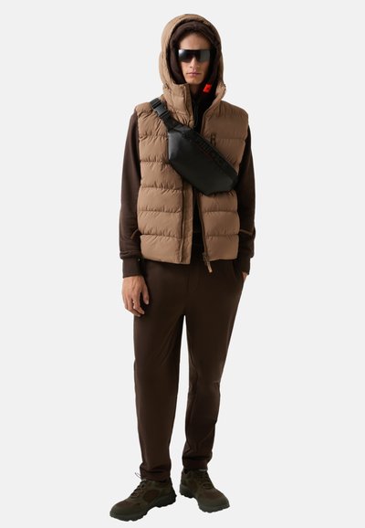 Braune, puffy Weste über einem braunen Hoodie, braune Hose und dunkelgrüne Schuhe. Schwarze Umhängetasche mit Logo und Sonnenbrille. Lässiger Outdoor-Stil.