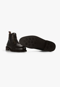 Botas de tobillo de cuero negro con una superficie texturizada, cordones en la parte delantera y punta redonda. La suela presenta patrones de tracción circulares para un mejor agarre.
