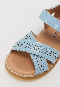 Sandal bleu clair en cuir verni avec des motifs floraux découpés, des bords festonnés et une semelle en cuir marron. Sangle ajustable incluse.