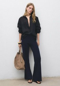 Donna che indossa una camicia nera oversize e pantaloni a zampa d'elefante blu, che tiene una grande borsa tote in suede beige, in piedi contro un muro bianco testurizzato.