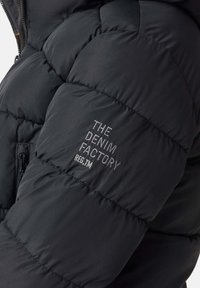 Svart pufferjacka med quiltat mönster. Har dragkedja och broderad text "THE DENIM FACTORY" på ärmen. Mjuk textur, isolerad.