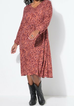 Femme portant une robe midi à manches longues orange et violette à motifs, avec des bottes de cowboy noires, debout devant un fond blanc.