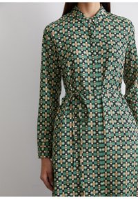 Abito verde a fantasia con forme geometriche in beige, nero e bianco, dotato di colletto e cintura in vita. Tessuto liscio con una brillantezza.