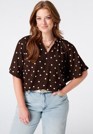 Femme aux longs cheveux ondulés porte un haut noir à manches courtes avec des pois roses et un jean bleu clair, souriante avec une main dans la poche.