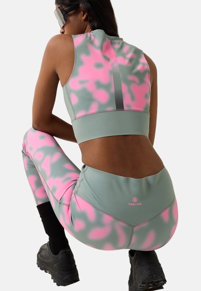 Frau hockt in grauem und pinkem Camouflage-Sportoutfit mit schwarzen Stiefeln und futuristischem Visier, von hinten vor schlichtem Hintergrund gezeigt.