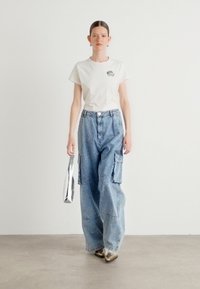 White cotton t-shirt with small logo, light blue denim cargo pants with side pockets, and metallic silver bag. Brown heeled shoes.