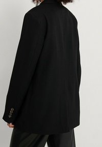 Blazer nero con una vestibilità rilassata, caratterizzato da colletto a rever, spacco singolo e polsini a quattro bottoni. Texture liscia con design su misura.