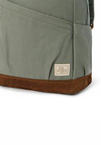 Vista laterale di uno zaino khaki con fondo in suede marrone e etichetta beige del marchio DaKine cucita vicino alla base.