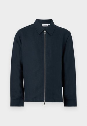 Mężczyzna noszący okrągłe okulary przeciwsłoneczne, granatową kurtkę, białą i granatową koszulę w paski, beżowe spodnie z szerokimi nogawkami oraz białe trampki, stoi na białym tle.