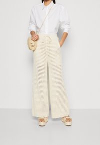 Szerokie spodnie szydełkowe w kolorze kremowym, z teksturowanym otwartym splotem, talią ze sznurkiem i falbankowanym dołem. Zestawione z białą koszulą na guziki.