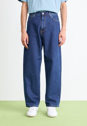 Personne portant un jean en denim bleu foncé ample, une chemise bleu clair et des baskets blanches, debout devant un fond uni.