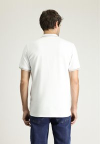 Biała koszulka polo z krótkim rękawem, wykonana z teksturowanej tkaniny, z subtelnymi złotymi akcentami na kołnierzyku i brzegach rękawów. Noszona z niebieskimi dżinsami.