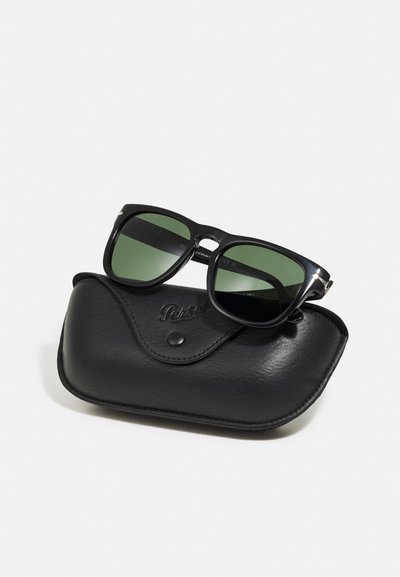 Black sunglasses with green lenses, featuring a square frame and silver accents, resting on a textured black leather case.