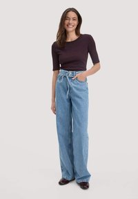 Donkerpaarse aansluitende top en lichtblauwe high-waisted wijde spijkerbroek met een trekkoord. Model staat met één hand in de zak en glimlacht.