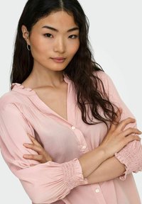 Lys rosa, knap-op skjorte med flæsekrave og ærmer med elastiske manchetter. Blødt stof med en glat tekstur, tætsiddende liv og afslappet pasform.