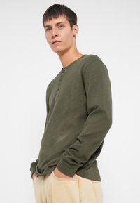 Dugih rukava, maslinasto zelena henley majica od teksturirane tkanine. Ima okrugli izrez s tri gumba i opušteni kroj.