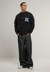 Svart kavaj med V-ringning och fem knappar. Har en blå "K"-applikation med stjärnor. Kombineras med löst sittande mörk denimjeans och vita sneakers.