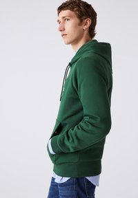 Joven con sudadera verde, con las manos en el bolsillo delantero, llevando una camisa de cuadros debajo y jeans azules, de pie de lado contra un fondo blanco.
