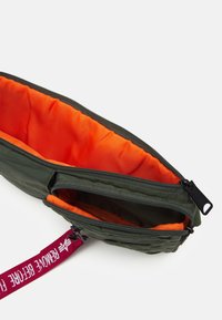 Sacchetto di tessuto verde con un interno arancione; presenta un compartimento con zip e una tracolla viola con testo stampato.