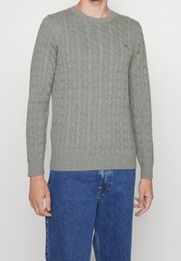 Šedý svetr s kabelovým vzorem, kulatým výstřihem a žebrovanými manžetami, kombinovaný s modrými džínami, s malým logem na hrudi.