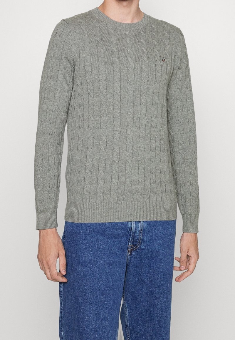Šedý svetr s kabelovým vzorem, kulatým výstřihem a žebrovanými manžetami, kombinovaný s modrými džínami, s malým logem na hrudi.