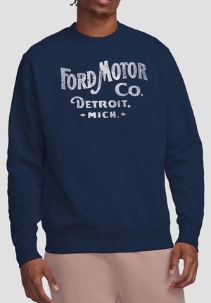 Man wearing navy blue sweatshirt with white "Ford Motor Co. Detroit, Mich." text printed on front, paired with light beige pants.