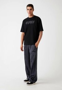 Fekete pamut póló szürke "GUESS" logóval, szürke melegítőnadrággal, amelyen szintén a "GUESS" logó található. A modell fekete tornacipőt visel.