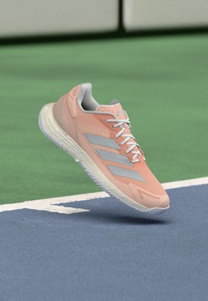 Lys rosa treningssko med grå detaljer og mesh-overdel, med hvit såle og teksturerte detaljer, plassert over en blå baneflate.