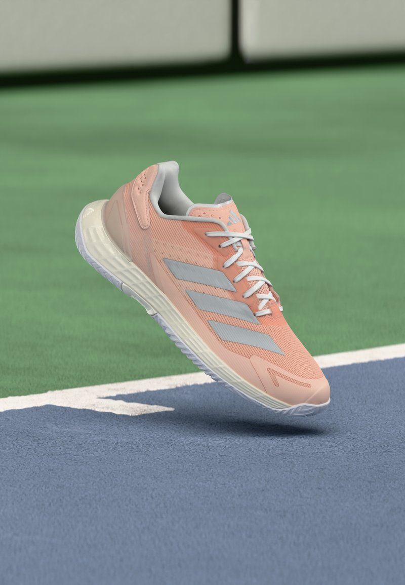 Svetloružová športová obuv so sivými akcentmi a vrchnou časťou z oka, vybavená bielou podrážkou a textúrovanými detailmi, umiestnená nad modrým povrchom kurtu.