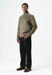 Olivgrüne Wildlederjacke mit hohem Kragen und Frontreißverschluss, kombiniert mit schwarzen Jeanshosen und braunen Schuhen. Schlichtes Design, strukturierte Oberfläche.