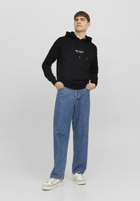Zwarte hoodie met grote voorzak, koord in de capuchon en witte logo-tekst. Gecombineerd met losse blauwe jeans en beige sneakers.
