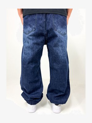 Jeanși baggy din denim de culoare albastru închis, cu o textură decolorată, având două buzunare pe spate și manșete elastice la gleznă. Asortați cu adidași albi.