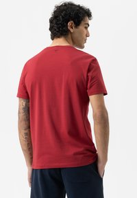 T-shirt en coton rouge à manches courtes, avec un col rond et un petit logo au dos. Le vêtement a une texture lisse.