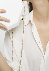 Bílá osmiúhelníková kabelka s texturovaným povrchem, zlatým řetízkovým popruhem a geometrickým zlatým akcentem. Nositá k světlé blůze.