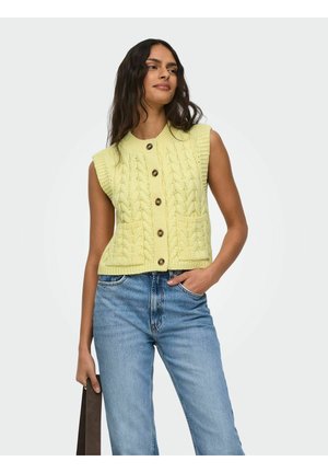 Femme portant un gilet boutonné en maille torsadée jaune pâle sans manches avec poches et un jean taille haute bleu, tenant une sangle de sac marron foncé.