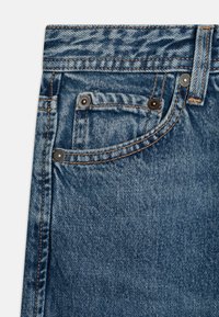 Közepes kék mosású farmernadrág réz színű szerelvényekkel, öt zsebbel és kontrasztos varrással a varratok mentén.