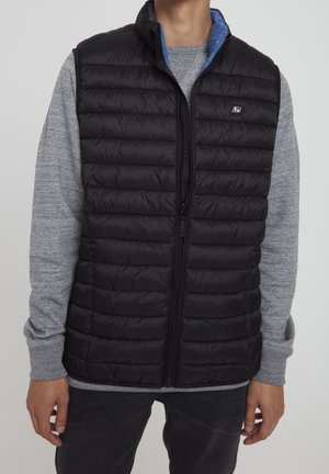 Man wearing black quilted vest over long-sleeve gray shirt with blue inner collar, standing against plain gray background.