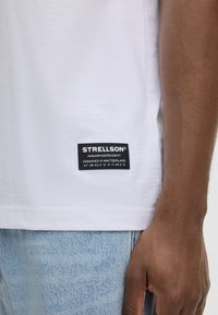 Weißes Baumwoll-T-Shirt mit einem schwarzen Markenlabel am linken Saum. Kombiniert mit hellblauer Denim, zeigt es eine weiche Textur und klassischen Schnitt.