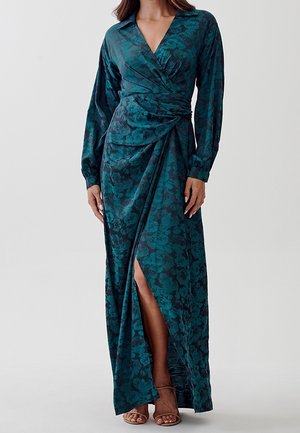 Femme portant une longue robe portefeuille fleurie bleu sarcelle et noire à manches longues avec une fente haute sur la cuisse, associée à des talons ouverts couleur camel.