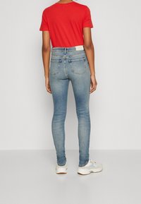 Vaqueros ajustados en denim azul claro con una cintura alta, con bolsillos traseros y una etiqueta en la cinturilla, combinados con una camiseta roja y zapatillas blancas.
