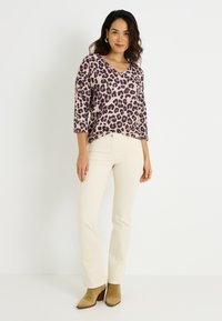 Suéter de cuello en V con estampado de leopardo púrpura, mangas de tres cuartos y pantalones acampanados beige. La modelo lleva botas tobilleras color arena, de pie contra un fondo blanco.