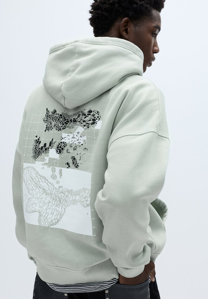 Lichtgroene hoodie met een grafische afbeelding op de rug met zwart-witte illustraties, een rasterpatroon en een gestructureerde afwerking.