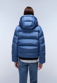 Giacca a vento blu con cappuccio, caratterizzata da strisce orizzontali trapuntate. Il tessuto esterno è lucido e liscio, abbinato a jeans chiari.